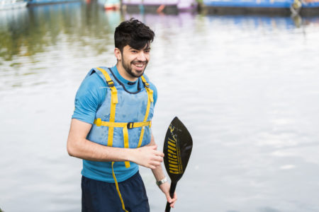 A man wearing a life jacket carries a canoe paddle in front of a body of water