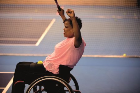 A female in a wheelchair celebrating a successful shot during an indoor tennis game.