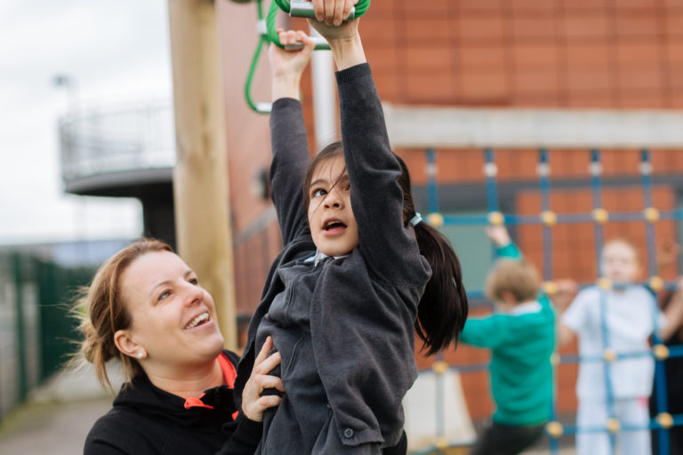 Child at school hanging from monkey bars with help from teacher